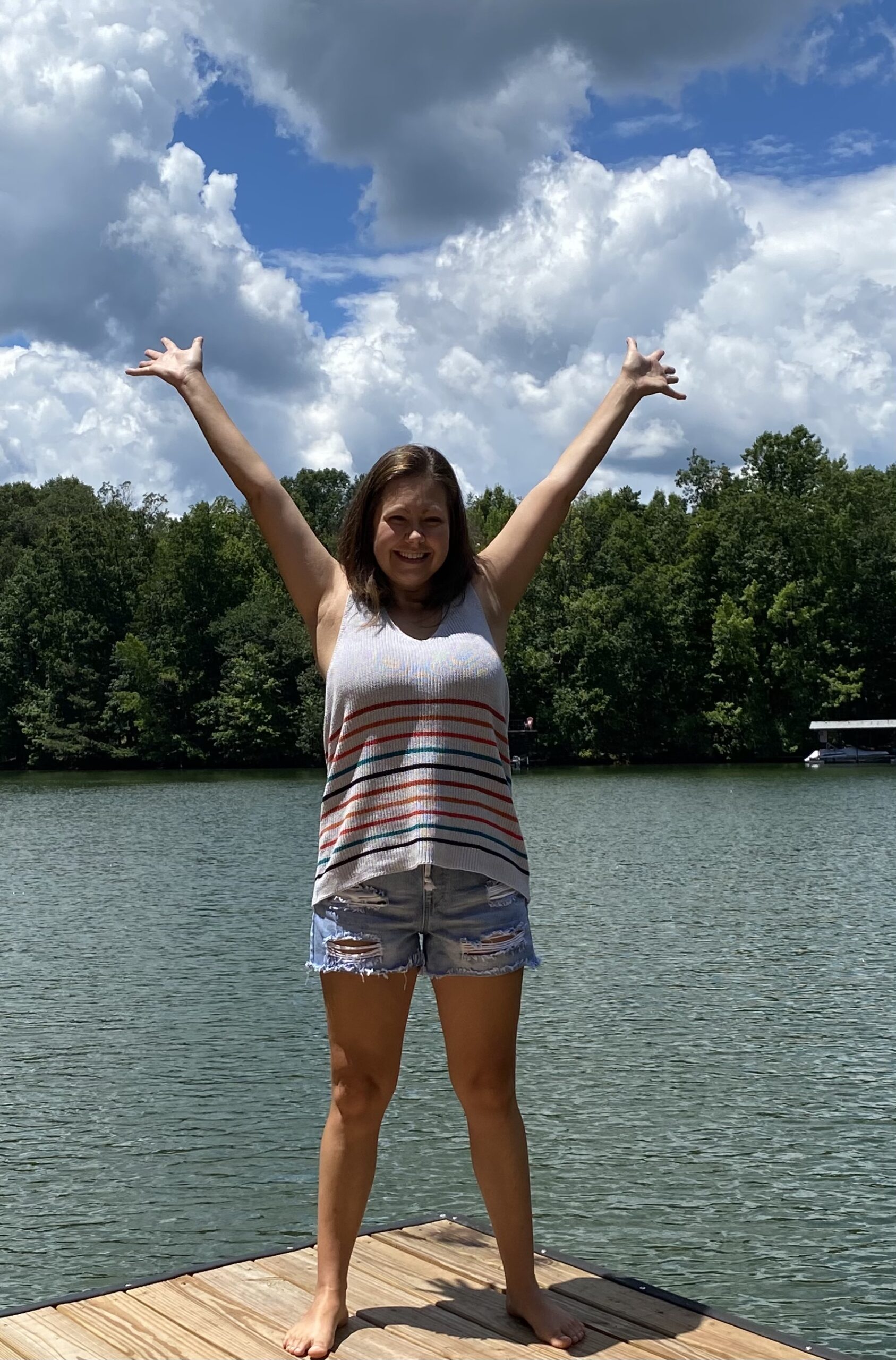 woman with a gray tank top and jean shorts, standing on a dock on a lake, with her arms up in the air in a V shape, smiling, feeling free