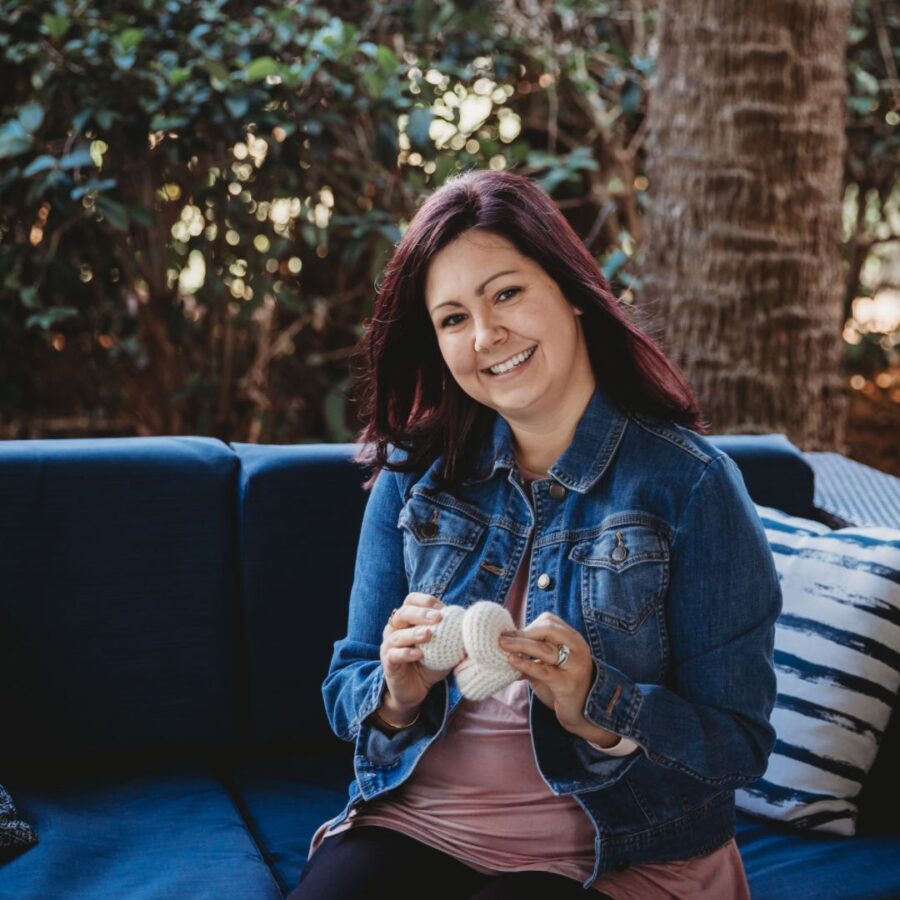 white woman with jean jacket and light pink shirt, holding a crochet breast and mouth, smiling and demonstrating a wide open gape of the crochet mouth to get a good latch on the crochet breast