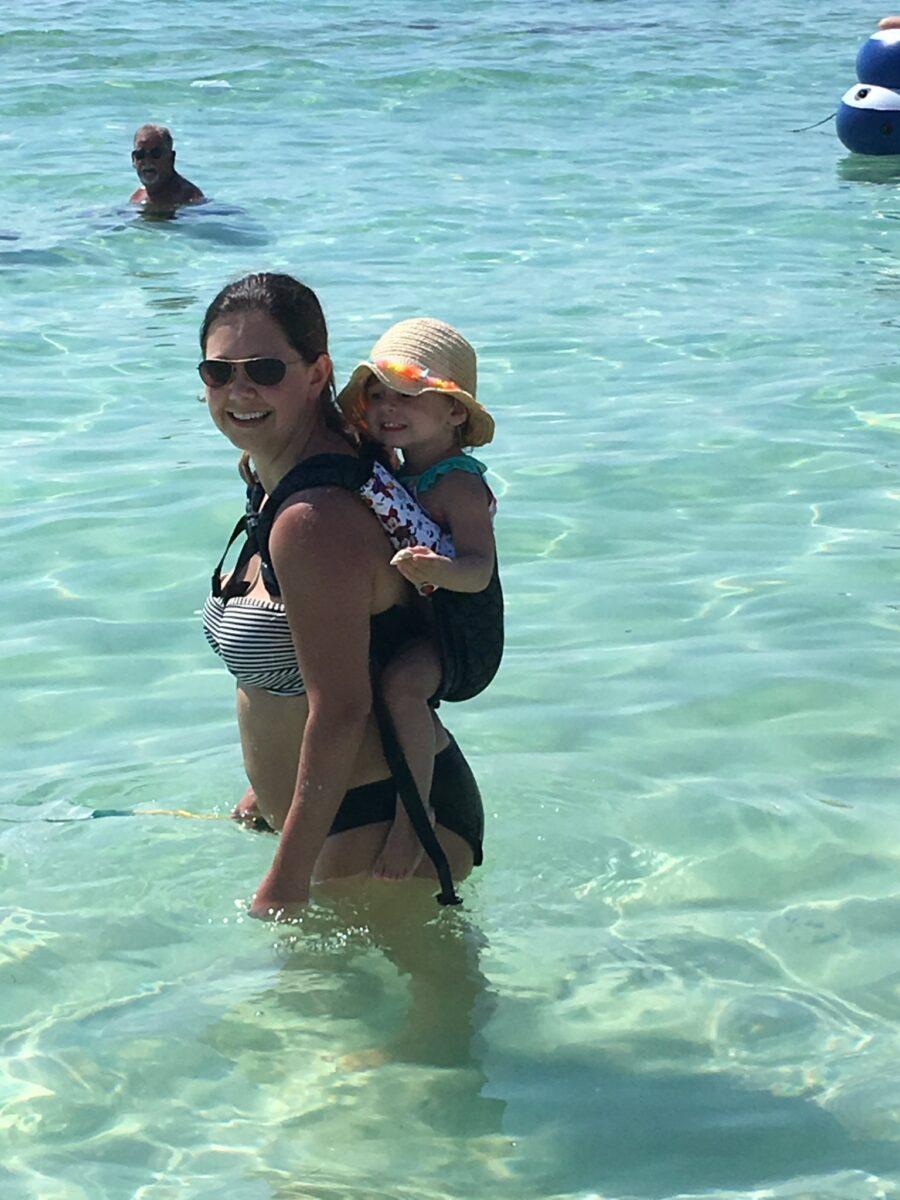 babywearing mom with toddler on her back, in a hat, in the beautiful blue green ocean waters