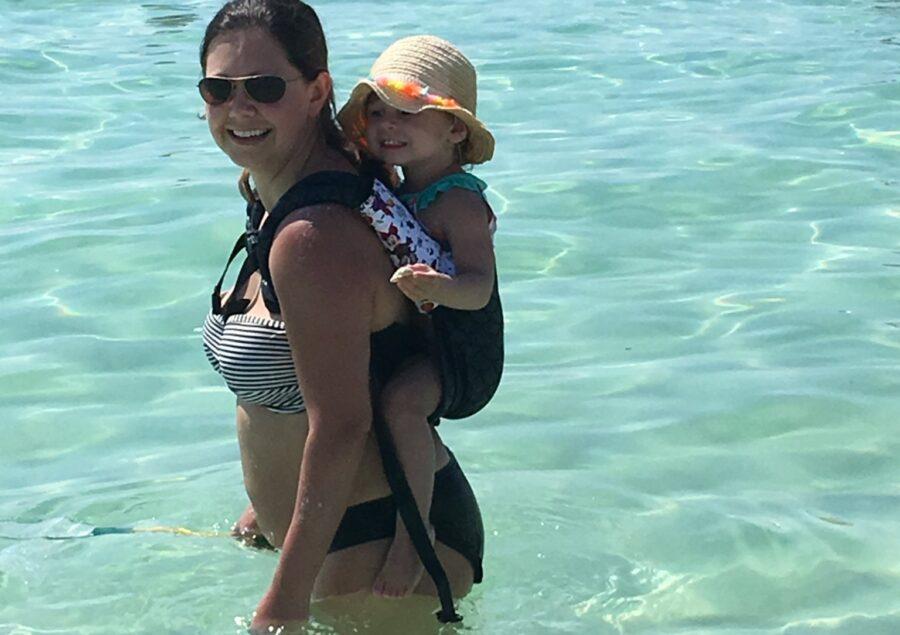 babywearing mom with toddler on her back, in a hat, in the beautiful blue green ocean waters